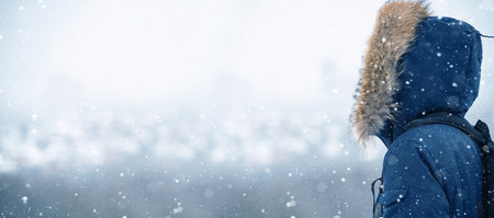 Back view of traveler in a blue jacket with a fur hood and a backpack on the background of a winter landscape during snowfallの写真素材