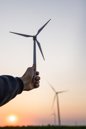 Man holds toy wind turbine in his hand. Real wind turbines in the background.の写真素材