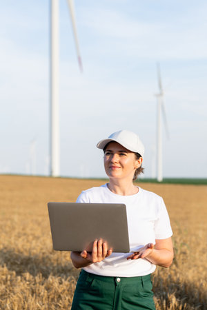 Woman farmer wearing white cap and t-shirt with laptop. Wind turbines in the background..の写真素材