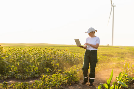 Woman farmer wearing white cap and t-shirt with laptop. Wind turbines in the background..の写真素材