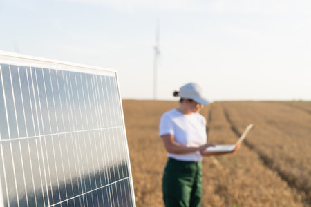 Woman farmer wearing white cap and t-shirt with laptop stands next to solar panel. Wind turbines in the background..の写真素材