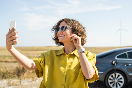 Woman with curve hairs wearing yellow shirt and sunglasses stands next to charging electric car and holds smartphone. Wind turbines in the background..の写真素材
