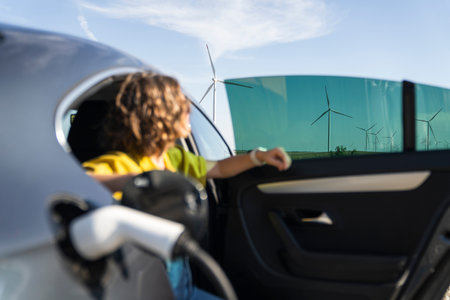 Woman with curve hairs wearing yellow shirt sits into charging electric car. Wind turbines in the...