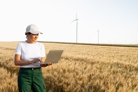 Woman farmer wearing white cap and t-shirt with laptop. Wind turbines in the background..の写真素材