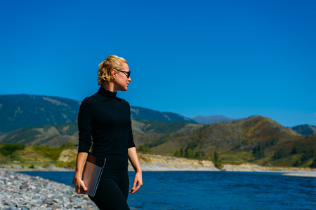 Freelancer with laptop on the nature. Blogger hipster traveler business lady pretty girl   finished work outdoor. Young blondy woman with sunglasses in black standing next to mountain riverの写真素材