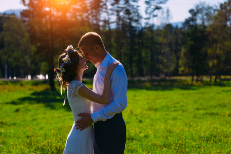 Wedding shot of the adorable newlyweds softly kissing on the meadow at the background of the trees during sunset.の写真素材