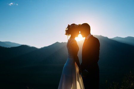 Happy wedding couple staying and kissing over the beautiful landscape with mountains during sunsetの写真素材