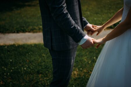 Wedding theme, holding hands newlyweds on green grass background. Hands of the bride and groom close-up. A couple on their wedding day, a touching moment.の写真素材