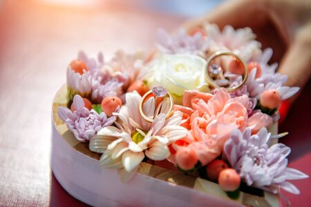 Wedding rings lie among fresh flowers in small wooden box, close up. Gold wedding rings on flower...
