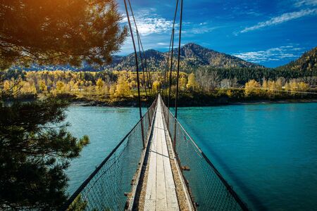 Pedestrian suspended wooden bridge over mountain river. Long bridge across the river against background of mountains covered with forest, golden autumn, indian summer warm sunny day.の写真素材