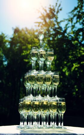Pyramid of champagne glasses stands on table outdoor. Sparkling drink in the glasses on green foliage background. Welcome drink at wedding celebration, anniversary. Vertical image.の写真素材