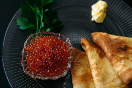 Traditional thin Russian pancakes with red caviar, butter and greens on a black wooden background. の写真素材