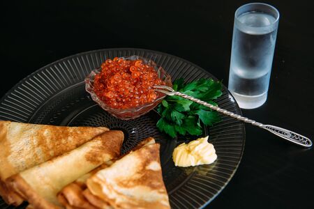 Golden russian pancakes with red caviar and butter in transparent plate near the glass of ice vodka isolated on black background. Restaurant of national cuisine.の写真素材