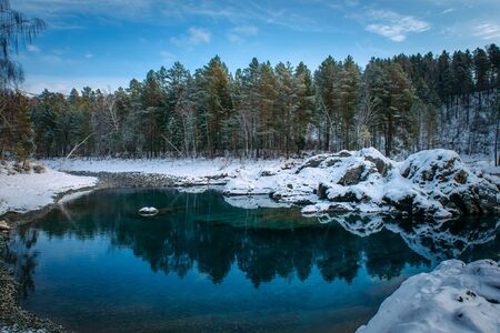 Winter panorama, small turquoise lake in the mountains among snow-covered forest. Trees are reflected in the lake water. Majestic nature of Altai mountains. の写真素材