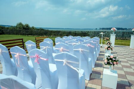 Exit registration of the newlyweds, wedding ceremony under open sky. Seating guests overlooking the river. Rows of chairs with white capes, wedding arch, floral design.の写真素材