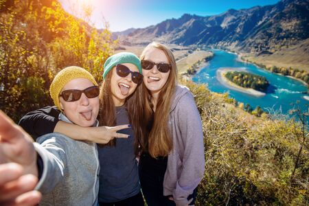 European women in sunglasses laugh doing selfie against mountain background. Young female hikers...