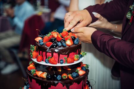 Luxury wedding cake decorated with chocolate, icing, fresh berries. Newlywed cut big beautiful cake at a wedding banquet, close-up.の写真素材