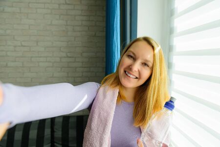 Young woman doing selfies after workout. Smiling attractive fitness girl with towel and bottle of water after training. Sport at home during lockdown.の写真素材