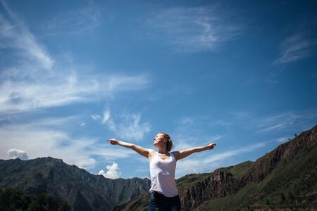 Happy female traveller in a white t-shirt against the beautiful mountains and blue sky on sunny day. Cheerful young girl enjoying a trip in the Altai mountains.の写真素材