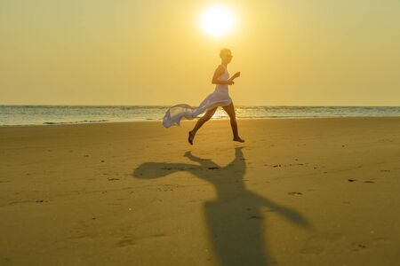 A happy young caucasian blonde woman in sunglasses and a fluttering white dress runs down the beach at sunset. Concept of recreation at sea and a healthy lifestyle.の写真素材