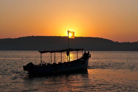 Picturesque sunset in the sea bay. The sun shines through the flag on a pleasure boat. Concept of leisure and tourism. Sea excursions in Goa. Boat in the rays of setting sun.の写真素材