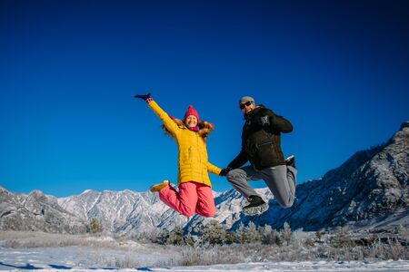 Young cheerful couple in bright clothes jump on the background of snowy mountains on winter sunny day. Guy and girl have fun during new year holidays in the mountains. Christmas and winter activities.の写真素材
