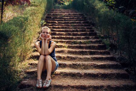 Cute woman in denim shorts sitting on step of the stairs in summer park. Stylish blonde on a blurry background. Beautiful young girl in a pensive mood.の写真素材