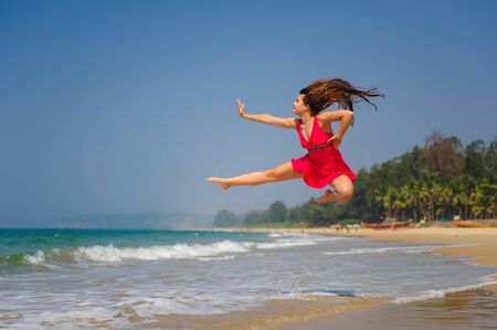 Happy young caucasian woman jumping high above wet sand in tropical sea on sunny day. Barefoot, a sporty girl in short red dress jumps and makes figures in the air.の写真素材