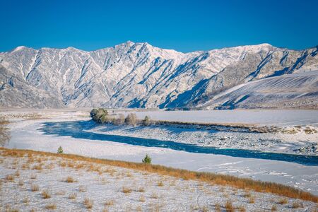 Majestic panoramic view of the valley covered with snow, turquoise river on the background of snow-capped mountains and clear blue sky. Sunny winter day in the mountains.の写真素材