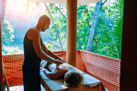 Slender cute male masseur does back massage to a girl outdoors. Young woman lies on massage table on a balcony overlooking palm greenery during wellness body massage session. Therapy at home.の写真素材
