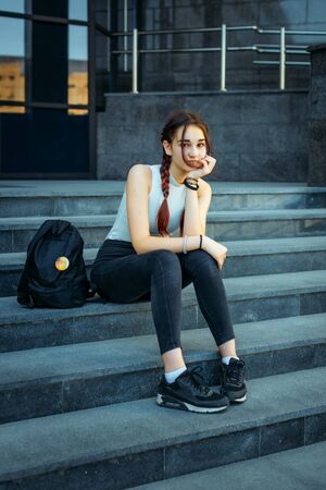 Beautiful European student with a backpack sitting on the steps near the University, close-up. A young pretty student in jeans and sneakers resting after class.の写真素材