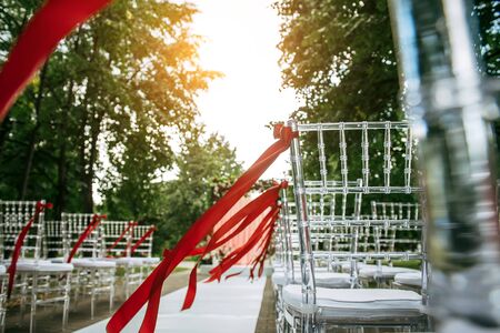Transparent stylish chairs decorated with red ribbons before the wedding ceremony outdoor. Rows of chairs in the park, abstract background.の写真素材