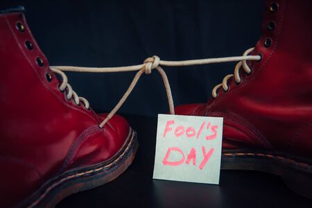 Stylish red shoes with laces tied together on black background. Boots covered by sticky note with text. April fool's day concept.の写真素材