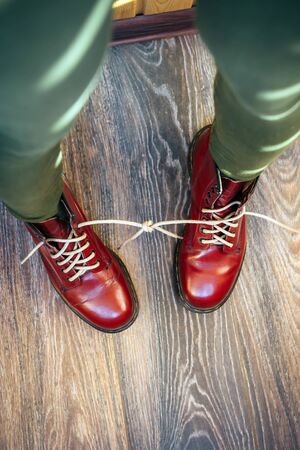 Female legs in stylish bright red boots with thick white laces tied together on wooden background, top view. Woman in funny pants and big size shoes. April Fools' day, pranks and fun.の写真素材