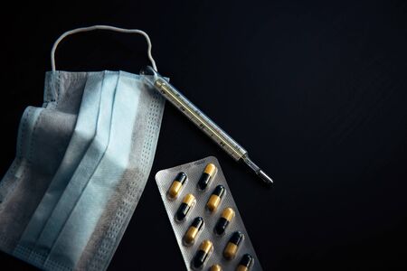 Medical face mask, thermometer and capsules in blister pack, close-up. Thermometer and medicine on black background. Epidemic season, coronavirus, influenza, acute respiratory diseases.の写真素材