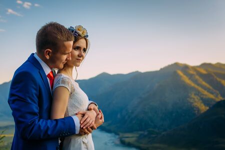 Young newly wed couple, bride and groom kissing, hugging on perfect view of mountains, river and...