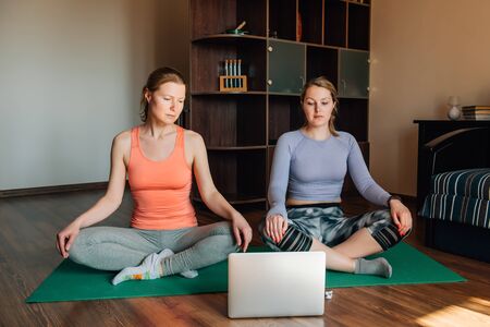 Two attractive young women doing stretching, yoga online at home. Self-isolation is beneficial, self-development and education on Internet. Healthy lifestyle concept.の写真素材