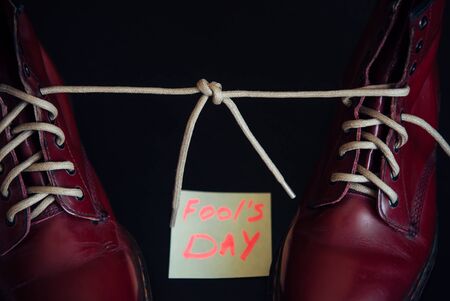 Stylish red shoes with laces tied together on black background. Boots covered by sticky note with text. April fool's day concept.の写真素材