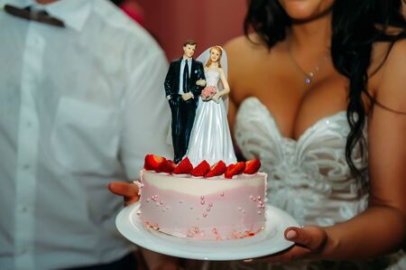 Wedding cake in the hands of bride in a white dress, close-up. Beautiful cake with strawberries decorated with figures of newlyweds.の写真素材