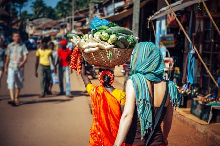An Indian woman in red Sari carries a large basket of vegetables on her head. Tourists and locals in India. A crowded street in Gokarna, Karnataka.の写真素材