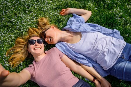 Two young happy woman doing selfie on green grass outdoor. Loving female couple lying face to face on the flowering meadow and holding hands. Serenity, youth, unisex love, friendship.の写真素材