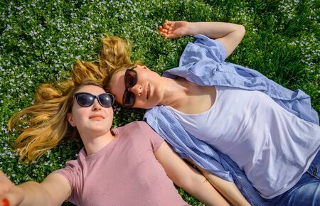 Two young happy woman doing selfie on green grass outdoor. Loving female couple lying face to face on the flowering meadow and holding hands. Serenity, youth, unisex love, friendship.の写真素材