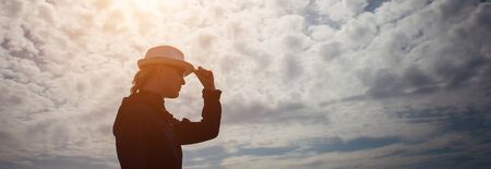 Female silhouette in profile with hat in hand against the mysterious sky with clouds. Outline of woman in a hat illuminated by the sun, copy space.の写真素材