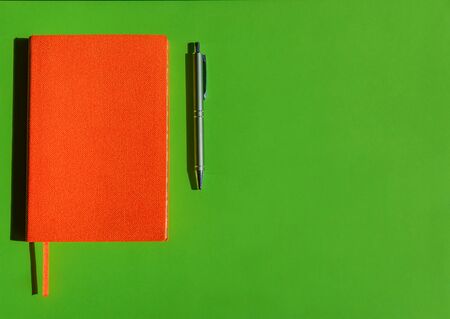 Bright orange notepad and stylish grey pen on green background with copy space. Concept of office products, diaries.の写真素材