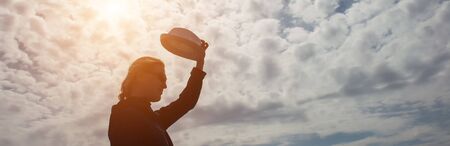 Female silhouette in profile with hat in hand against the mysterious sky with clouds. Outline of woman in a hat illuminated by the sun, copy space.の写真素材