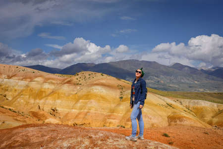 Young woman in jeans and sunglasses stands against beautiful mountains and blue sky on sunny day....
