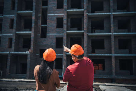 Rear view builders a man and a woman in orange helmets stand on the background of a brick apartment...