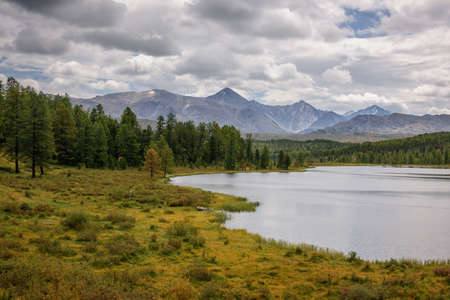 Quiet lake among the mountain peaks and pine forests, cloudy sky. Idyllic panorama of autumn, Alpine...