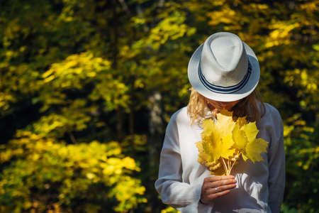 Beautiful young woman in white clothes and hat tilted her head to the bouquet of yellow leaves in her hands. Elegant blonde, face hidden by hat. Freedom, love, happiness. Indian summer, golden autumn.の写真素材