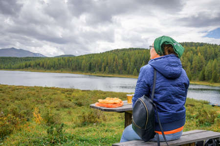 Young woman traveler sitting back to the camera and admiring the beautiful view of mountains. Wooden table and bench for tourists rest near lake. Pause for snacks and relax.の写真素材